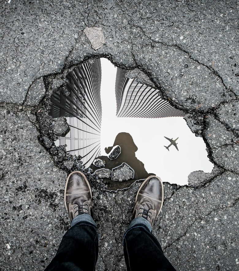 A person standing over a broken mirror embedded in pavement, reflecting a building and plane.